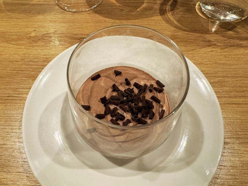 A rich chocolate mousse topped with dark chocolate shavings, elegantly served in a clear glass bowl on a white plate, ready to be enjoyed on the Costa Smeralda cruise ship.