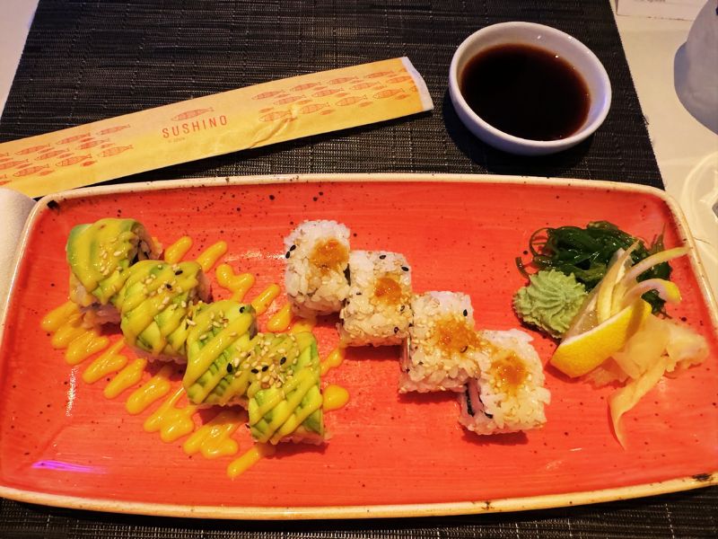 An artfully arranged sushi platter on a vibrant orange plate from Sushino aboard Costa Smeralda, featuring avocado rolls with a mango sauce drizzle and uramaki topped with orange fish roe, served with soy sauce and a side of pickled ginger and seaweed salad.