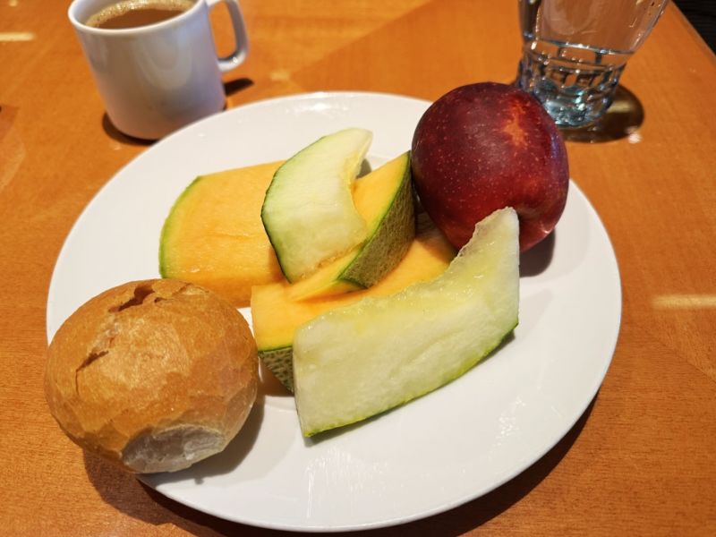 A healthy breakfast plate with melon and honeydew slices, a fresh apple, and a whole grain bread roll, accompanied by a cup of coffee and a glass of water on a wooden table aboard the Costa Smeralda cruise ship.