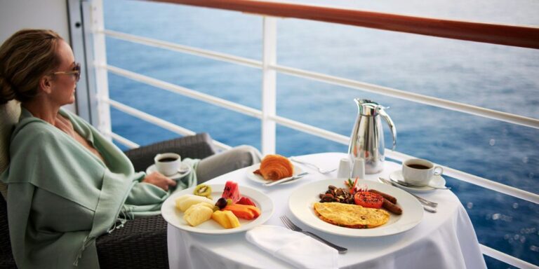 This image portrays a tranquil breakfast scene on the balcony of an Oceania Cruises stateroom. A woman in a comfortable robe is seated, gazing out at the ocean, with a well-presented breakfast spread before her. The table holds a variety of breakfast items including a plate of fresh, sliced fruits, a croissant, and a hot dish with an omelet, grilled tomato, and sausages, accompanied by coffee and juice. This setting captures the essence of luxurious cruising, offering a private and serene dining experience with a breathtaking ocean view.