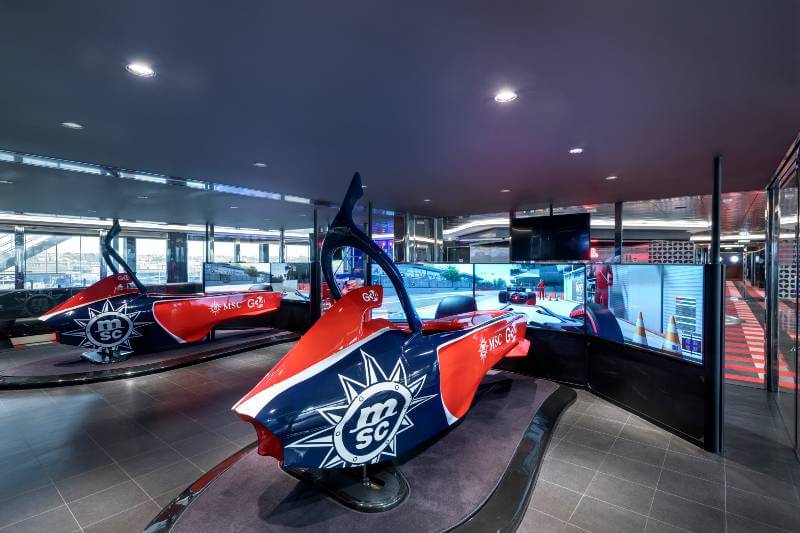 A high-tech Formula One simulator on an MSC cruise ship, featuring racing car replicas in MSC livery against a backdrop of large screens simulating a thrilling racetrack experience, offering an adventurous activity at sea.