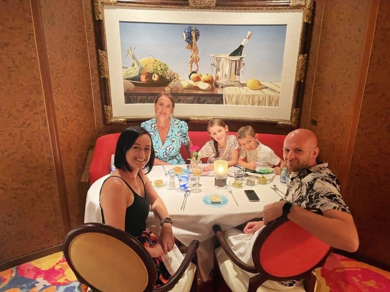 Cruise Mummy and her family are enjoying a meal at a specialty dining restaurant on the Fred. Olsen Bolette cruise ship. They are seated at a round table adorned with a candle and elegant place settings. The backdrop features an ornate framed painting of a still life with fruit and champagne. The family, dressed in vibrant, summery clothing, is smiling and engaged in conversation, creating a warm and pleasant dining atmosphere.