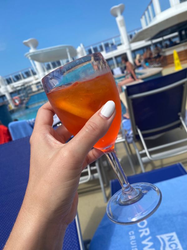 A close-up of a hand with white nail polish holding an Aperol Spritz on the sun-drenched deck of an NCL cruise, with poolside loungers and the ship's structure in the background, capturing the 'Free at Sea' promotion's essence of relaxation and enjoyment.