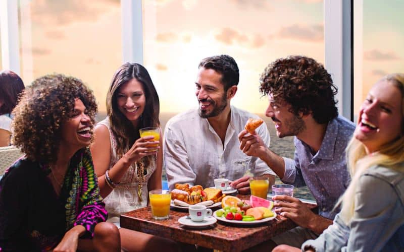 A vibrant group of friends enjoying breakfast together, with fresh fruit, pastries, and orange juice on the table, in a brightly lit room with a window showcasing a sunset or sunrise. Their laughter and the casual, cozy atmosphere suggest a happy start to the day.