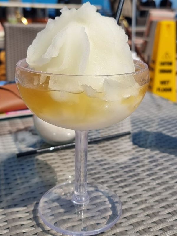 A Frozen 24K Golden Margarita on a Princess cruise ship, served in a wide-rimmed glass with a mound of yellow slush. The drink is placed on a woven table, capturing a sunny, outdoor setting with blurred background elements, including a "Wet Floor" sign and other passengers, creating a refreshing and luxurious atmosphere.