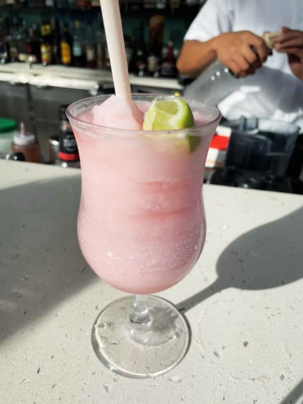 A Frozen Watermelon Margarita on a cruise ship, served in a hurricane glass with a lime wedge garnish and a straw. The drink is placed on a bar counter, with a bartender preparing drinks in the background, creating a refreshing and tropical setting for enjoying this pink slushy cocktail.