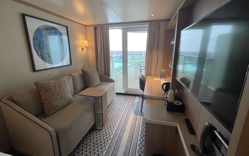 A tastefully decorated P&O Cruises Iona Balcony Cabin featuring two twin beds with gold runners and a geometric-patterned carpet. The room is accessorized with round mirrors, soft lighting, and has a cozy sitting area by the balcony door, inviting guests to relax with a view of the ocean.