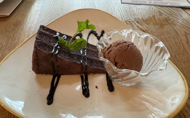 A slice of rich chocolate cake drizzled with syrup and a sprig of mint, accompanied by a scoop of chocolate ice cream in a clear glass dish, artistically plated on a ceramic plate, offering a tempting dessert option at Center Parcs.