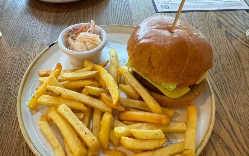 A classic burger with cheese and lettuce on a brioche bun, served with a side of golden fries and coleslaw in a small dish, presented on a ceramic plate with a rustic wooden table background, capturing the essence of a hearty meal at Center Parcs.