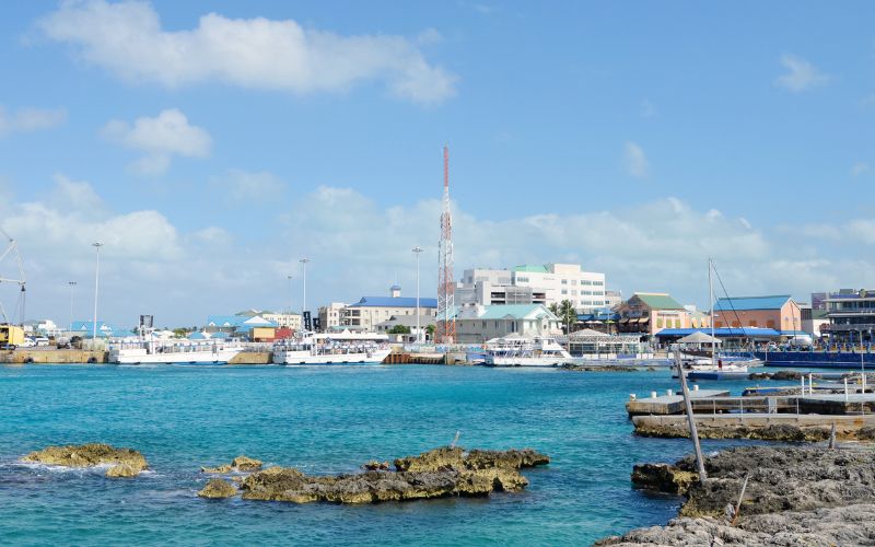 The vibrant coastal setting of Georgetown, Grand Cayman, featuring clear turquoise waters and a lively waterfront lined with a variety of buildings. The scene includes modern commercial structures, recreational boats docked along the pier, and natural rocky outcrops at the shoreline. The clear sky and bright sunshine enhance the inviting tropical ambiance of the capital city, showcasing its blend of natural beauty and urban development.