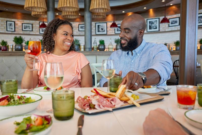 A couple shares a joyous moment at Giovanni's on Royal Caribbean's Freedom of the Seas, with the woman holding a refreshing Aperol Spritz and the man engaging in a delightful culinary experience, dipping bread into olive oil. The backdrop of the restaurant's chic interior, along with their assortment of salads and charcuterie, sets the scene for a luxurious dining experience at sea.