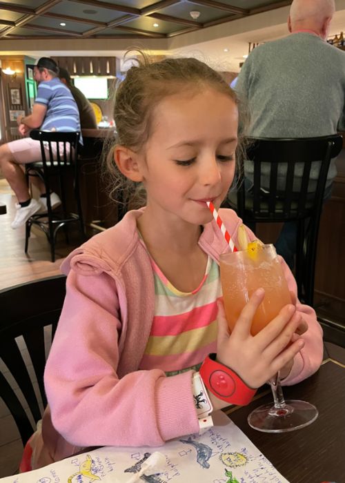 My daughter enjoying a fruity drink with a striped straw, dressed in a striped top and pink hoodie, seated in a casual dining setting on MSC Virtuosa with patrons in the background.