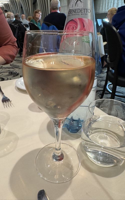 A close-up of a glass of chilled rosé wine on a dining table, with a bottle of Benedetto natural mineral water in the background. The setting suggests a refined dining experience on a cruise, with other passengers visible in the softly lit restaurant.