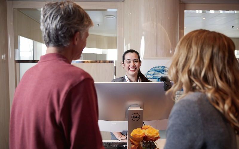 A smiling customer service representative assists two passengers at the Guest Services Priority Line for Club Orange on a Holland America Line cruise ship, with a bouquet of orange roses on the counter.