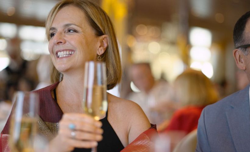 A joyful female guest on a Holland America Line cruise ship raises a glass of wine in a toast, smiling brightly in a semi-formal dining room filled with natural light and other passengers.