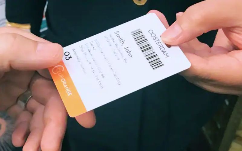 Hands holding a Holland America Line Club Orange keycard with the name 'Smith John' printed on it, highlighting the exclusive access and personalization for Club Orange members aboard the Oosterdam.