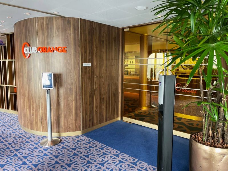 The Club Orange Restaurant entrance on the Rotterdam, a Holland America Line cruise ship, with its distinctive logo on a wooden backdrop, a hand sanitizing station, and a decorative plant, welcoming passengers to an upscale dining environment.