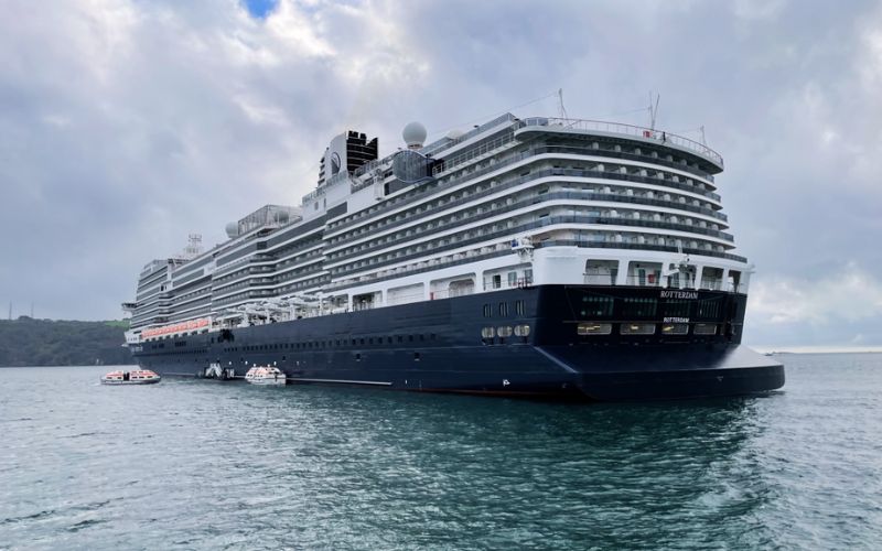 The MS Rotterdam, a Holland America Line cruise ship, anchored at sea with lifeboats at the ready, showcasing its multi-deck structure and distinctive navy blue hull on an overcast day.