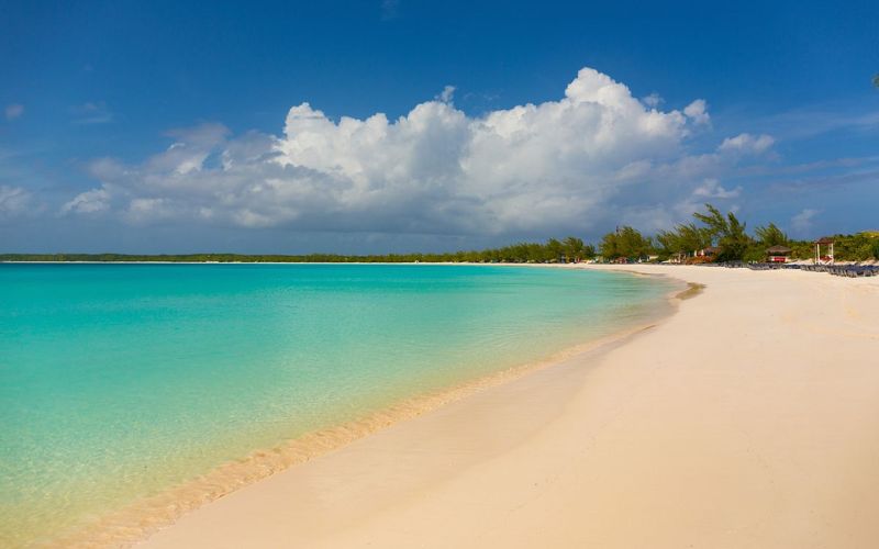 The serene coastline of Half Moon Cay, with its pristine white sand and clear, shallow turquoise waters, extends towards a tranquil horizon under a dramatic sky. The few scattered beachgoers and lush greenery in the distance add to the island's peaceful and untouched atmosphere.