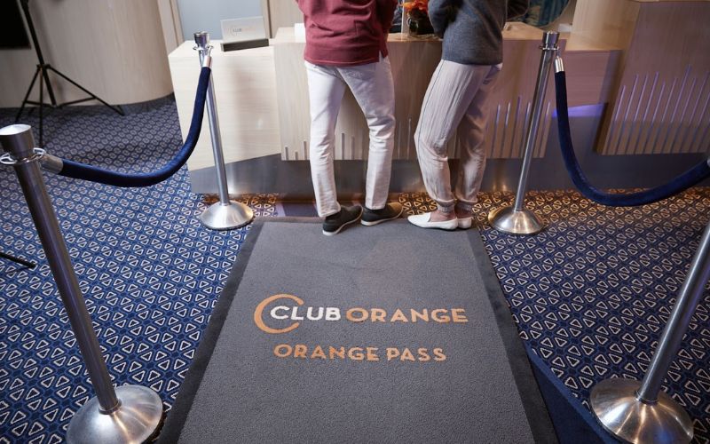 Priority check-in area for Club Orange on a Holland America cruise, showcasing two guests from the waist down standing on a grey mat with 'CLUB ORANGE ORANGE PASS' text, flanked by blue velvet ropes and silver stanchions.