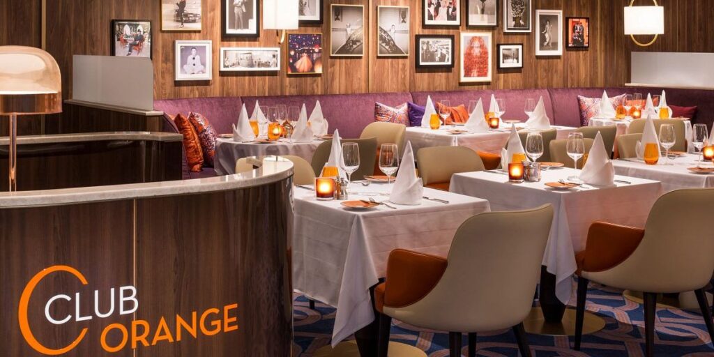 Interior view of Club Orange restaurant on a Holland America cruise ship, featuring a modern dining area with white-clothed tables set for dinner, orange and purple accent pillows on the booths, and walls adorned with framed photographs.