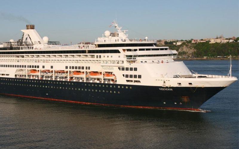 The Holland America Veendam cruise ship, featuring its dark hull and white superstructure, glides by a coastline bathed in the warm glow of the setting sun.