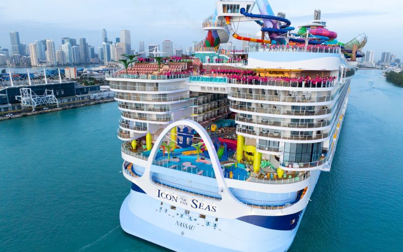 Aerial view of the 'ICON of the SEAS' cruise ship, docked in a harbor with the city skyline in the background. The ship is adorned with colorful waterslides and an elaborate aqua park visible on the upper decks, highlighting its family-friendly amenities.