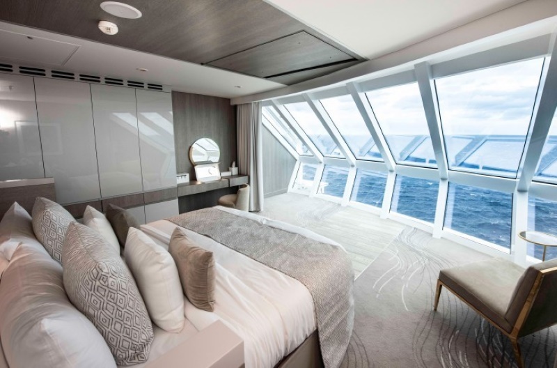 The luxurious Iconic Suite on a Celebrity cruise ship, featuring a king-sized bed with plush pillows, a sleek design with neutral tones, and an array of panoramic windows offering breathtaking, uninterrupted ocean views.