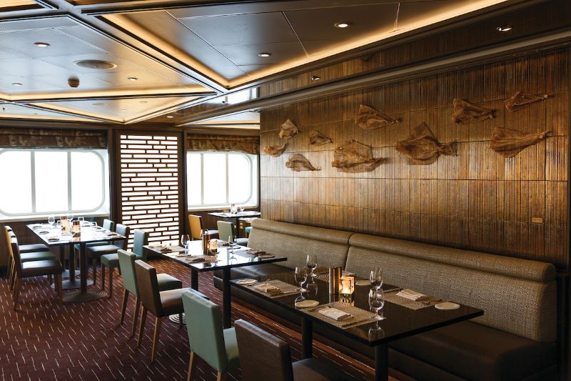 The Indochine restaurant on a Silversea cruise ship, capturing the serene dining atmosphere with plush seating, tables set with fine crystal, and a wall adorned with wooden fish sculptures, complemented by natural light from porthole windows.