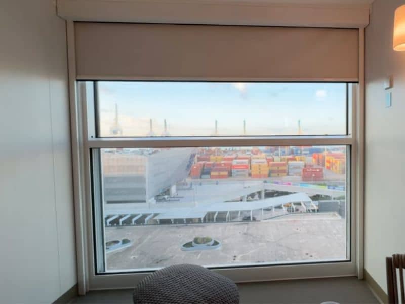 View from the 'Infinite Balcony' aboard Royal Caribbean's 'Icon of the Seas,' showing a bustling port scene. The large window frames a view of colorful cargo containers and the port infrastructure, blending the indoor comfort with the industrial exterior. A subtle, wicker chair is partially visible, enhancing the room's connection to the outdoor scenery.