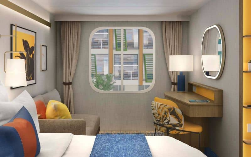 Interior view of another stateroom aboard Royal Caribbean's 'Icon of the Seas,' showcasing modern amenities and décor. The room features a plush bed with blue and white bedding, colorful accent pillows, a beige sofa, and a small work desk with an oval mirror. A large porthole-style window provides a view of the ship's exterior passageways, adding natural light to the space.