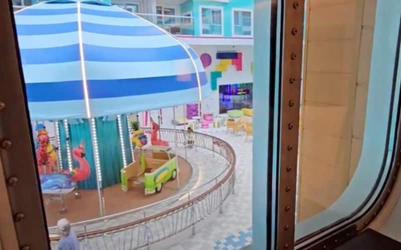 View from the window of an interior stateroom on Royal Caribbean's 'Icon of the Seas,' overlooking the ship's colorful and lively Surfside neighborhood. The window frames a bustling children's area featuring a whimsical carousel under a large blue and white striped umbrella, surrounded by vibrant playground equipment and seating areas.