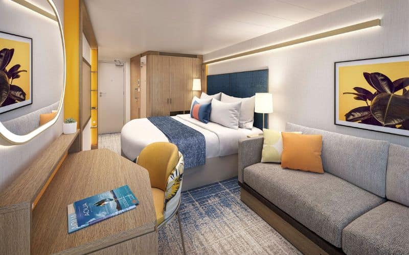 Interior view of a stateroom on Royal Caribbean's 'Icon of the Seas' featuring a cozy arrangement. The room includes a round wooden table, a plush gray sofa, a comfortable bed with white and blue bedding, and colorful throw pillows. Artwork and mirrors decorate the walls, creating a bright and welcoming atmosphere.
