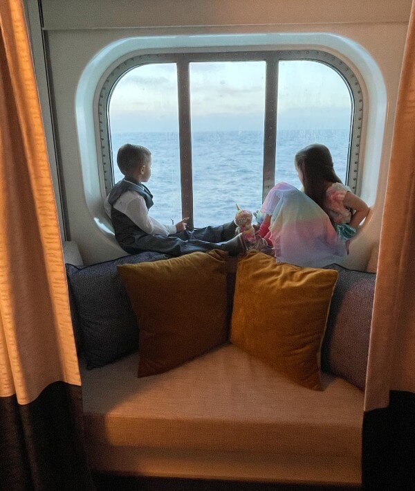 Two children are seated on a window seat inside P&O Iona cabin, gazing out at the ocean through a large sea view window. The boy is dressed in a vest and trousers, while the girl wears a colorful dress and holds a stuffed animal. The cozy seating area is adorned with plush cushions, providing a comfortable spot to enjoy the scenic view.