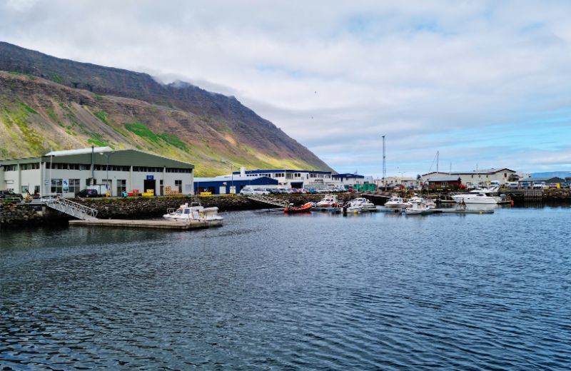 This image offers a picturesque view of Isafjordur, Iceland, showcasing the serene harbor filled with boats against a dramatic backdrop of steep mountains. The harbor area features functional buildings and warehouses, highlighting the town's fishing industry. The calm water reflects a cloudy sky, emphasizing the remote and tranquil atmosphere of this northern Icelandic town nestled within the deep fjords. The landscape conveys a sense of rugged beauty and isolation typical of Icelandic coastal communities.