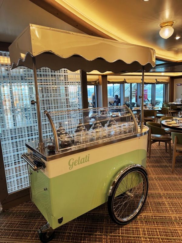 Onboard the Seabourn Venture, a charming Italian gelato cart adds a sweet touch to the dining options. This vintage-style cart, painted in soft pastel green with a beige canopy, is equipped with a glass cover revealing a tempting assortment of gelato flavors. Parked within the buffet area, it offers a delightful treat for guests, enhancing their culinary experience with a taste of Italy. The warm ambient lighting and chic backdrop of the dining area create a cozy atmosphere for enjoying this classic dessert.