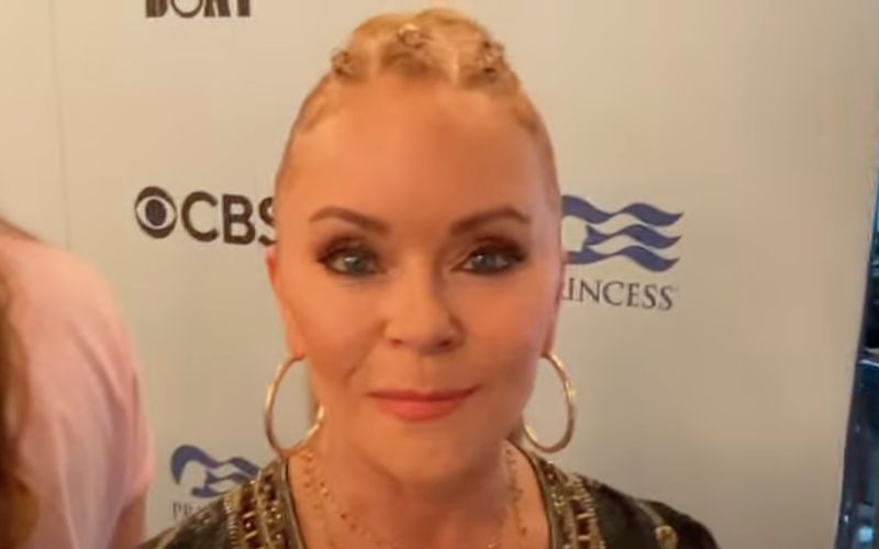 Jill Whelan with blonde hair styled in an updo, featuring large hoop earrings and a patterned garment. She is posing in front of a backdrop with the logos of CBS and Princess Cruises.