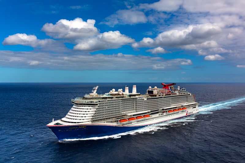 Carnival Jubilee cruise ship, with its distinctive red and blue funnel, cruises on a calm blue ocean under a partly cloudy sky, showcasing its multiple decks and the iconic water slide on the top deck.