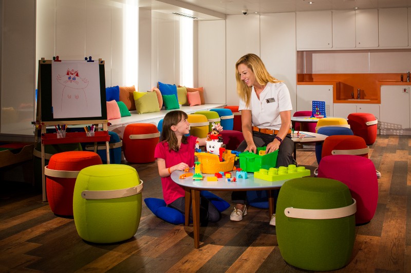 A cheerful scene in the Kid's Club on HAL MS Koningsdam with a young girl and a staff member engaging in play with colorful building blocks at a table, surrounded by vibrant, cozy seating.