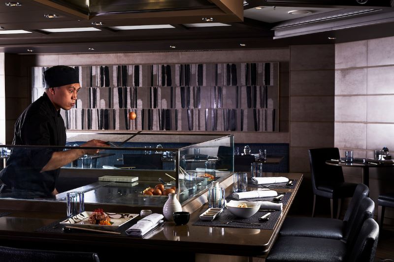 An attentive chef prepares sushi at the Kaiseki restaurant on a Silversea cruise ship, with a view of the elegant sushi bar setting featuring dark chairs, modern decor, and a serene ambiance.