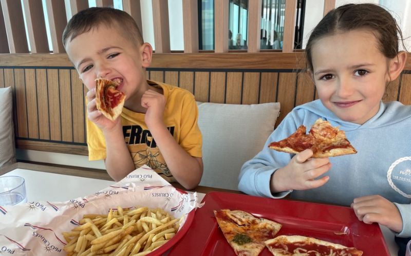 Two joyful children enjoying a meal of pizza and fries, with a boy playfully biting into a slice of pizza and a girl smiling while holding her slice, all set against a modern restaurant backdrop.