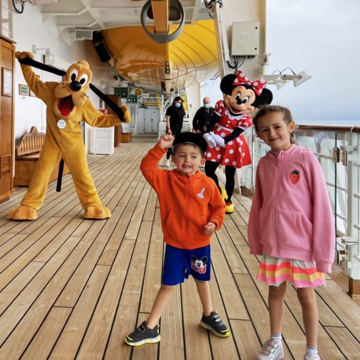 Two cheerful children posing on the deck of a Disney Cruise ship, with costumed Pluto and Minnie Mouse characters in the background, creating a memorable vacation moment.