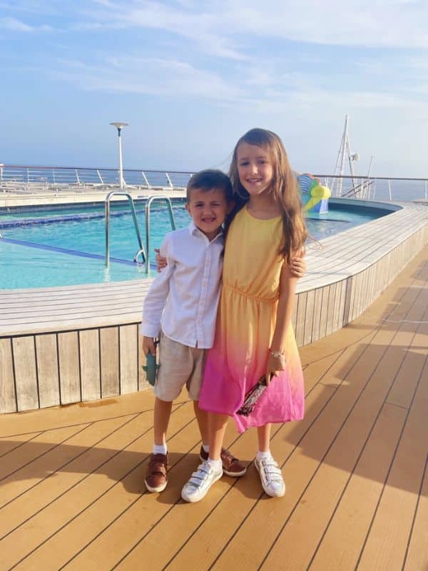 Two smiling children, a girl in a yellow ombre dress and a boy in a white shirt and beige shorts, stand together on the deck of a Fred. Olsen cruise ship, with the ship's pool and the open sky in the background.