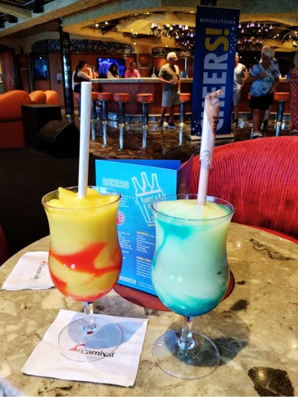 Two cocktails on a Carnival cruise ship: "Kiss On The Lips," a yellow and red layered drink, and "Twilight Zone," a blue and white swirled drink. They are served in hurricane glasses with straws, placed on a marble table with Carnival-branded napkins, against a backdrop of a bar area with people socializing and a sign promoting the beverage program.