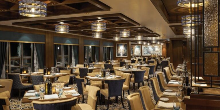 The sophisticated interior of La Cucina restaurant on the NCL Bliss, featuring a luxurious dining area with beige and navy blue chairs, elegantly set tables, and ornate circular chandeliers, creating an inviting atmosphere for guests.