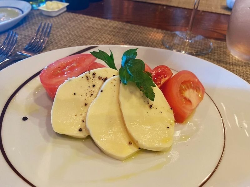 A classic Caprese salad with fresh slices of mozzarella cheese and ripe tomatoes, garnished with a sprig of basil, served on a white plate at La Cucina on NCL Getaway, embodying simple Italian elegance.