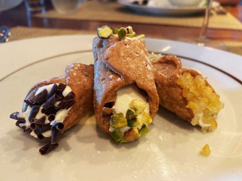 Delicious cannoli filled with creamy ricotta cheese, one garnished with chopped pistachios and the other with chocolate curls, presented on a white plate at La Cucina on NCL Getaway, offering a taste of Italy at sea.