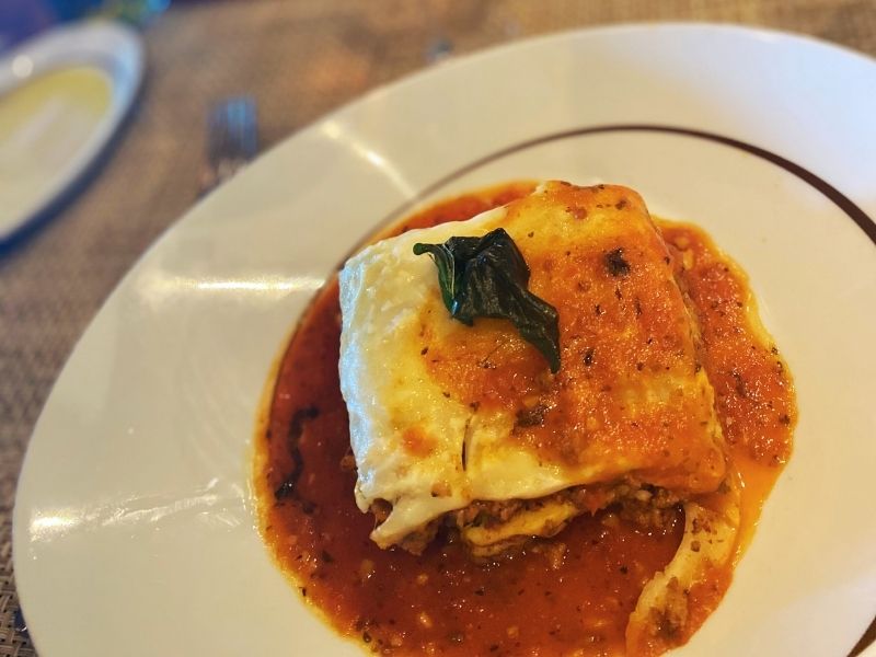 A savory portion of lasagna with molten cheese and a basil leaf garnish, bathed in a rich tomato sauce, served on a white plate at La Cucina on NCL Getaway, exemplifying classic Italian comfort food.