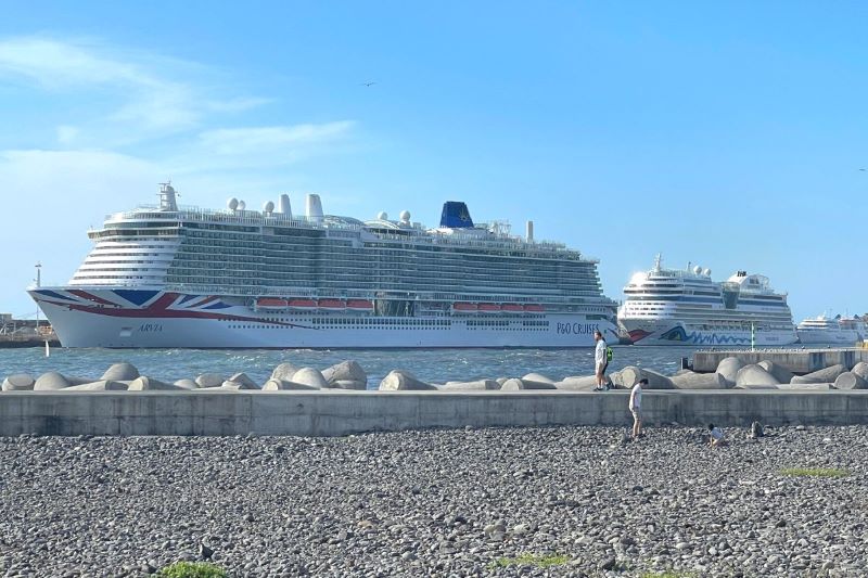 The image presents a clear day at the harbor with two imposing cruise ships in view. The closest ship, prominently displaying the Union Jack flag design on its hull, is identified as the P&O Arvia, a testament to its British heritage. In the background, another large cruise ship with a blue funnel is visible, both dwarfing the person strolling along the pebble beach in the foreground, showcasing the grand scale of these luxurious vessels.