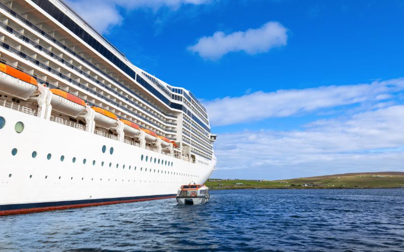 This image features a large cruise ship docked near the shores of Lerwick, Scotland, emphasizing the vast scale of the vessel against the rural landscape backdrop. A small tender boat is visible beside the cruise ship, ferrying passengers to and from the shore. The setting is serene with open waters and gentle hills in the distance under a clear blue sky, capturing the peaceful interaction between modern tourism and the natural environment of this Scottish locale.